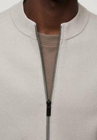 Chaqueta gris claro con cremallera, de tela texturizada y cuello acanalado. Presenta una cremallera en contraste y una capa interior sólida debajo.