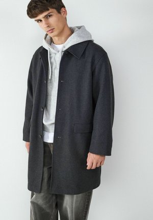 Donkergrijze wollen jas met een kraagontwerp, knoopsluiting en zijzakken, gelaagd over een lichtgrijze hoodie en gecombineerd met grijze broek.