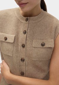 Beige mouwloze gebreide sweater met een ronde halslijn, met twee borstzakken en bruine knopen aan de voorkant. Zachte textuur.