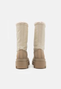 Korkeat beige nahkasaappaat, joissa on pörröinen vuori ylhäällä, paksut kumipohjat ja näkyvät saumat valkoista taustaa vasten.