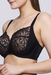 Sujetador negro con copas de encaje transparente y bordado floral, banda inferior sólida, tirantes ajustables y detalles de borde en forma de ondas.