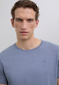 Junger Mann mit kurzen, lockigen Haaren, der ein schlichtes, hellblaues T-Shirt mit rundem Hals und einem kleinen Totenkopf-Logo auf der Brust trägt.