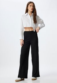 Zwarte wijde broek van textuurstof, gecombineerd met een witte cropped blouse met knopen. Model draagt casual witte sneakers.
