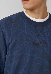 Primo piano di una persona che indossa un felpa blu navy con un sottile motivo di rametti e il testo "S.Oliver Elevated Since 1969" sul petto.