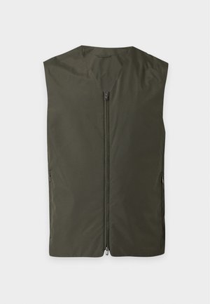 Homme portant une veste zippée vert foncé par-dessus une chemise marron à manches courtes, un jean large bleu clair et des baskets beige, debout avec une main dans la poche.