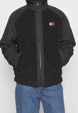 Uomo che indossa una giacca Tommy Jeans in pile nero e nylon con cerniera frontale e jeans di denim blu, mani nelle tasche, su sfondo uniforme.