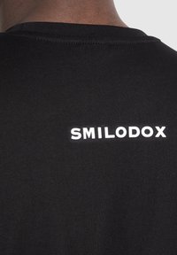 Musta t-paita pyöreällä kauluksella, jossa on valkoinen "SMILODOX" -logo painettuna selkäpuolelle, valmistettu sileästä puuvillasekoitekankaasta.
