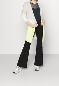 Hellbeige Cardigan mit gelben Akzenten, schwarzes Top mit Spitzenverzierungen und ausgestellte schwarze Hosen. Schuhwerk: weiße Sneaker mit blauen Akzenten.