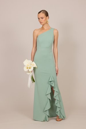 Femeie într-o rochie verde pal cu un umăr dezgolit și detaliu cu volane, ține crini albi calla, poartă cercei aurii și sandale pe fundal bej.