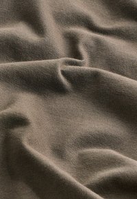 Hnědá bavlněná tkanina s měkkou, hladkou texturou a jemným elastickým strečem, vytvářející lehký a pružný vzhled s vlnícími se záhyby.