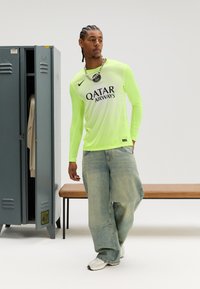 Neona dzeltens garroku sporta krekls ar svītrainu rakstu un melnu zīmolu. Valkāts kopā ar vaļīgām gaiši zilām džinsu biksēm un baltām sporta kurpēm.