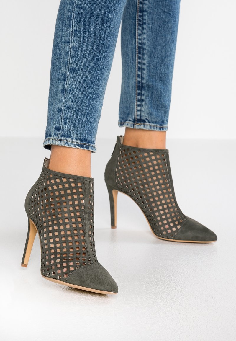 Botki na wysokim obcasie z ciemnozielonego zamszu, z geometrycznym wzorem wycięć i szpiczastym noskiem, w połączeniu z niebieskimi dżinsami.