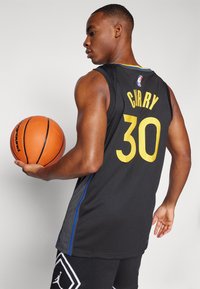 Sort basketballtrøje med gul "CURRY" og nummer "30" på ryggen; har blå accenter og et struktureret, åndbart stof. Holder en orange basketball.