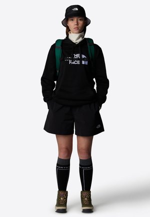 Mustapaita, jossa on "The North Face" -logo, mustat shortsit, vihreä reppu, musta ämpärihattu, harmaat polvisukat ja vihreät vaelluskengät.