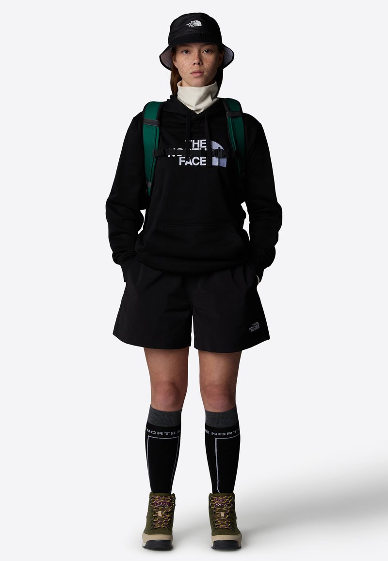 Sudadera negra con el logo de "The North Face", pantalones cortos negros, mochila verde, sombrero de cubo negro, calcetas grises hasta la rodilla y botas de senderismo verdes.