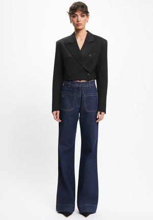 Młoda kobieta ubrana w krótką czarną dwurzędową marynarkę i ciemnoniebieskie, szerokie spodnie z wysokim stanem, stojąca na jednolitym tle.