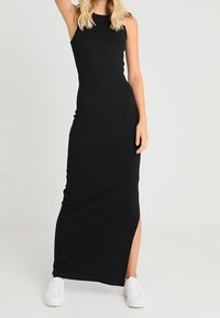 Mustapintainen ribattuja maksimekko, jossa on hihaton muotoilu, sivureikä ja tiukasti istuva siluetti, yhdistettynä valkoisiin tennareihin kontrastina.