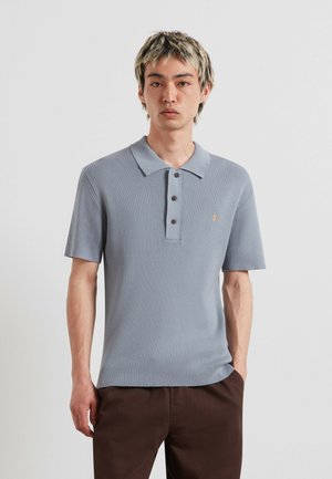 Giovane uomo con capelli corti tinti che indossa una polo azzurro chiaro a coste a maniche corte e pantaloni scuri, in piedi davanti a uno sfondo semplice.