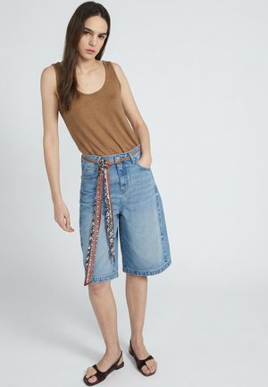 Jonge vrouw staat met één hand in haar zak, draagt een bruin mouwloos topje, knielange spijkershorts met een patroonriem en donkere sandalen.