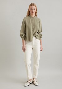 Olijfgroene button-up blouse met een mandarijnenkraag, opgeblazen mouwen en een ontspannen pasvorm, gecombineerd met witte rechttoe rechtaan jeans en casual schoenen.
