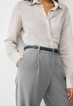 Frau trägt ein beigefarbenes, gestreiftes Hemd mit Knöpfen, das in eine hoch taillierte graue Hose gesteckt ist, dazu ein dunkelblauer Gürtel mit Kettengliedern.