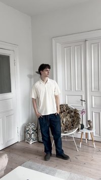 Camiseta polo blanca de manga corta con un bolsillo, combinada con pantalones de chándal azul oscuro con cordón. De pie en un suelo de madera cerca de muebles blancos.