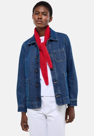 Femme portant une veste en denim bleu par-dessus une chemise blanche et un pantalon, accessoirisée d'une écharpe rouge en tricot nouée autour du cou.