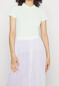 Valkoinen lyhythihainen polo-paita, jossa on kaulus ja nappilista, yhdistettynä laskostettuun hameeseen, jossa on violetteja raitoja. Sileä kankaan rakenne.