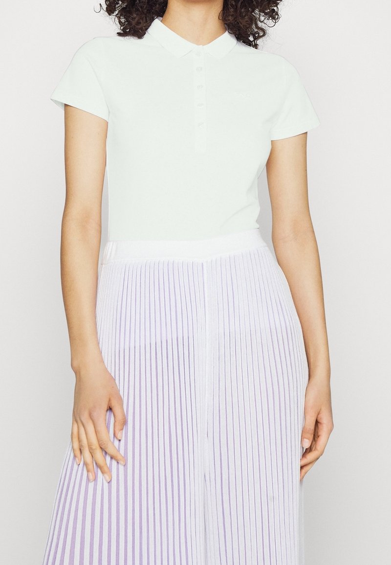 Valkoinen lyhythihainen polo-paita, jossa on kaulus ja nappilista, yhdistettynä laskostettuun hameeseen, jossa on violetteja raitoja. Sileä kankaan rakenne.