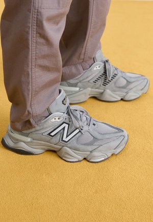 Personne portant des baskets New Balance grises et un pantalon marron, debout sur un sol recouvert d’un tapis jaune.