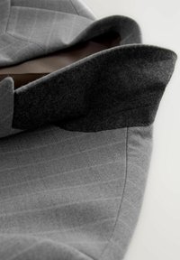 Abrigo de lana gris con una textura suave, que presenta un cuello doblado y un tejido oscuro contrastante a lo largo del borde interior. Patrón de raya diplomática sutilmente visible.