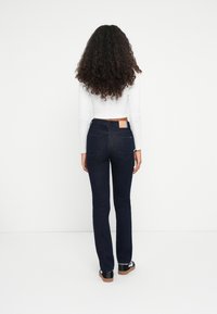 Jeans de mezclilla de color azul oscuro con corte acampanado, de talle medio, con bolsillos traseros y un parche de cuero marrón en la cintura. El modelo lleva una camiseta blanca de manga larga.