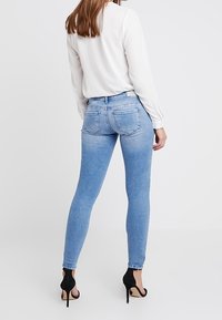 Jeans ajustados de color azul con un acabado desgastado, que presentan una cintura de altura media, bolsillos traseros y un corte slim. Combinados con una blusa blanca de manga larga y tacones.