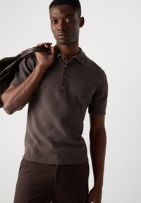 Camisa polo de punto acanalado marrón con mangas cortas, tapeta de tres botones y un acabado texturizado; modelo llevando un bolso oscuro sobre el hombro.