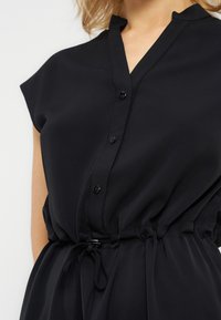 Γυναίκα που φοράει μαύρο φόρεμα με κουμπιά και κορδόνι στη μέση, με κοντά μανίκια, εμφανιζόμενη από τους ώμους έως το μέσο του κορμού.