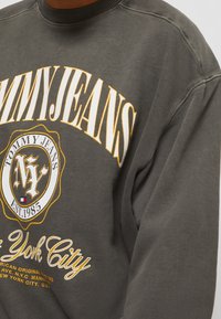 Felpa grigia scura con collo a girocollo a coste. Presenta una grande stampa "TOMMY JEANS" e un logo circolare con "NY" e "EST. 1985" in giallo e bianco.
