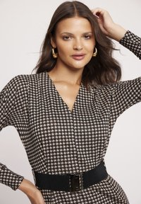 Sukienka w wzór houndstooth w odcieniach czerni i beżu, z długimi rękawami, dekoltem w kształcie litery V oraz szerokim czarnym elastycznym paskiem z prostokątną klamrą.