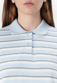 Close-up of a person’s collar and upper chest, wearing a light blue striped polo shirt with two buttons and subtle branding visible.