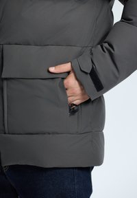 Gray puffer jacket with a textured surface, featuring a front pocket, Velcro closure on the sleeve, and an adjustable drawstring inside.
