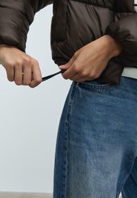 Ruke podešavaju uzicu na smešnom puffer jakni nosenoj preko plavih traperica protiv obične svetle pozadine.