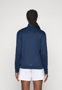 Person with braided hair in a ponytail wears a navy long-sleeve top and white shorts, standing facing away against a plain background.