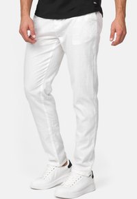 Pantalon en coton blanc avec une coupe slim et des poches latérales, associé à des baskets blanches avec des accents noirs au niveau du talon. Texture lisse, jambes droites.