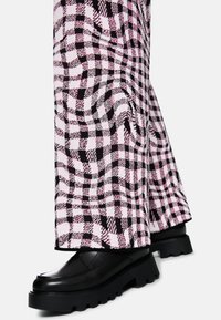 Schwarze klobige Schuhe, die mit rosa und schwarzen karierten weiten Hosen vor einem weißen Hintergrund getragen werden.
