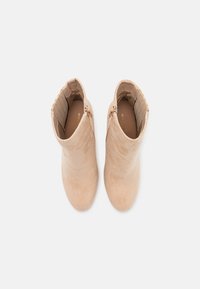 Bottines en daim beige avec un design élégant, bout pointu et zips latéraux. Doublure intérieure texturée avec le nom de la marque visible.