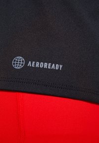 Černá sportovní tkanina s logem "AEROREADY" ve světlé šedé; červená textura pod ní má výrazné švy. Hladký povrch po celé ploše.