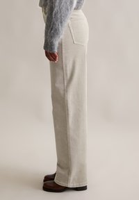 Beige Cordhosen mit weitem Bein, hoher Taille, Seitentaschen und einer strukturierten, gerippten Oberfläche. Braune Stiefeletten sichtbar.