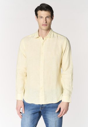 Uomo che indossa una camicia giallo chiaro a maniche lunghe con bottoni e jeans blu, in piedi davanti a uno sfondo chiaro e semplice.