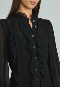 Blusa nera con colletto arricciato e pattina anteriore con bottoni decorativi. Il materiale è trasparente, creando una texture a strati.