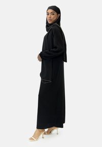 Schwarze Abaya mit langen Ärmeln und seitlichen Schlitzen, versehen mit goldenen Akzenten entlang der Ränder. Getragen mit einem passenden Hijab und beigen Absätzen.