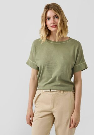 Blonde Frau trägt ein hellolivgrünes, kurzärmeliges Oberteil und beige Hosen mit einem geflochtenen Gürtel, steht vor einem schlichten grauen Hintergrund.